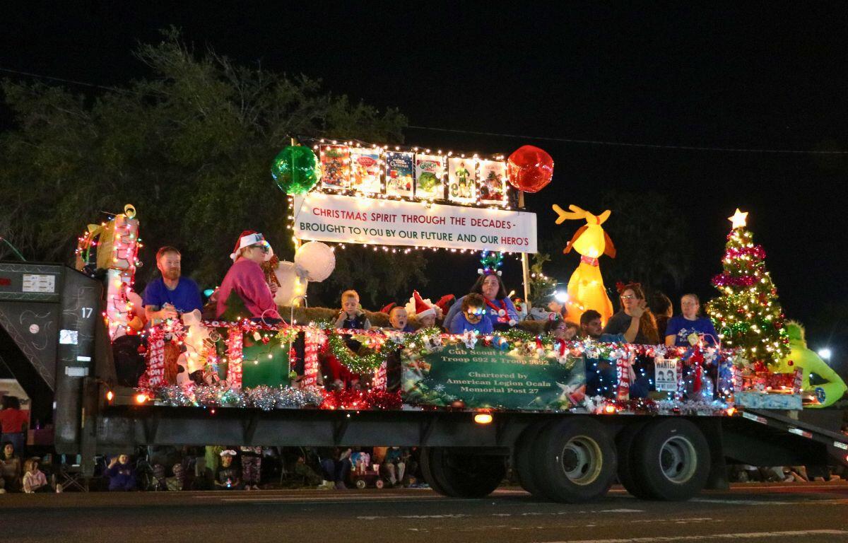 A truck lit by Christmas lights, inflatables and a tree drives down a dimly lit road with multiple people on top.