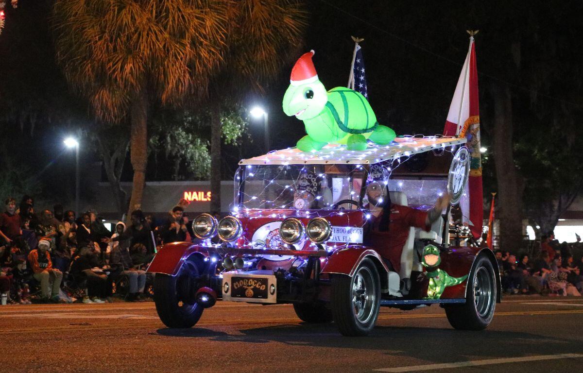 A small car has multiple Christmas lights, flags and an inflatable turtle with a Santa hat on top drives down a dimly lit road.