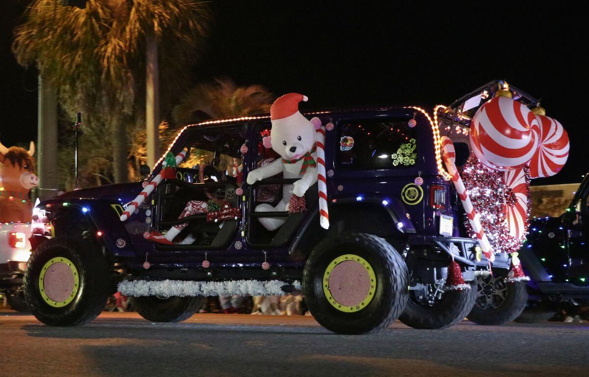 A jeep with multiple Christmas lights, candy cane decorations and an inflatable polar bear wearing a Santa hat hanging out of the back seat drives down a dimly lit road.