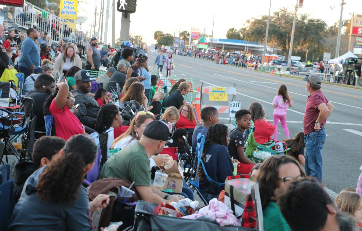 A crowd forms on the sides of a street with many in lawn chairs and some in stadium seating.