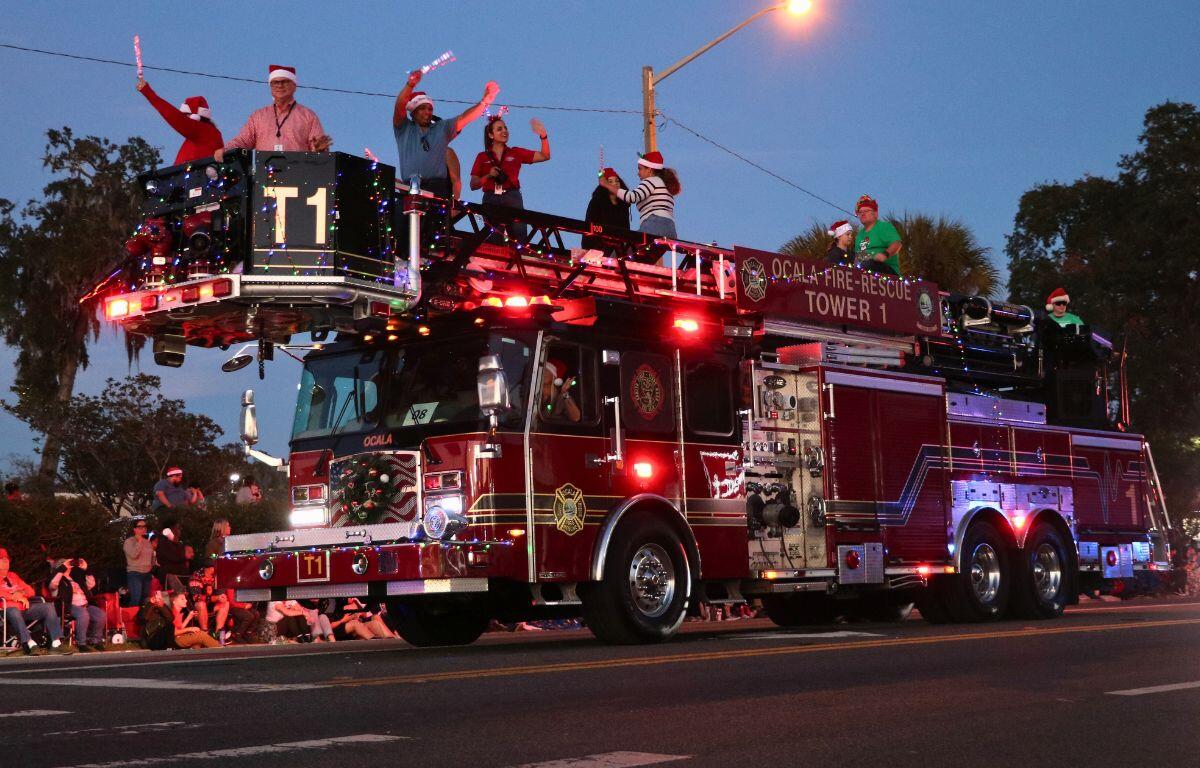 A large fire truck is decorated in Christmas lights, driving down a road while people in Santa hats wave to crowd from the top.