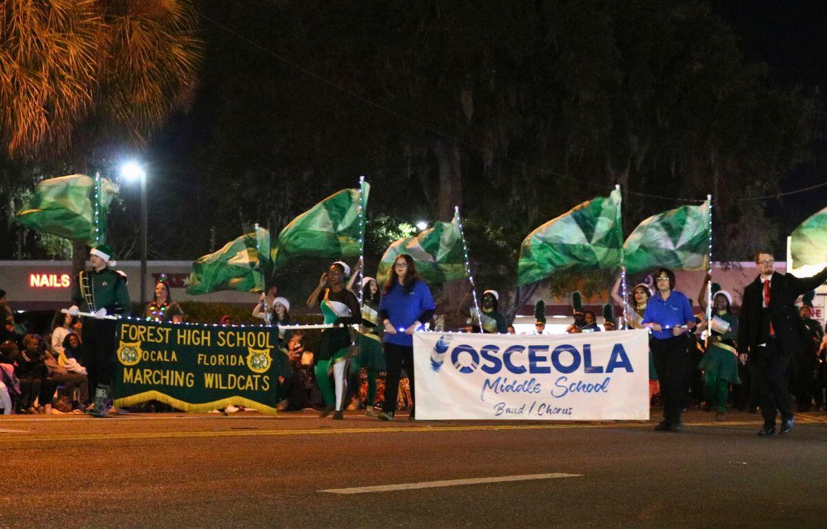 A group of students twirling flags and playing instruments march holding signs representing Forest High School and Osceola Middle School.