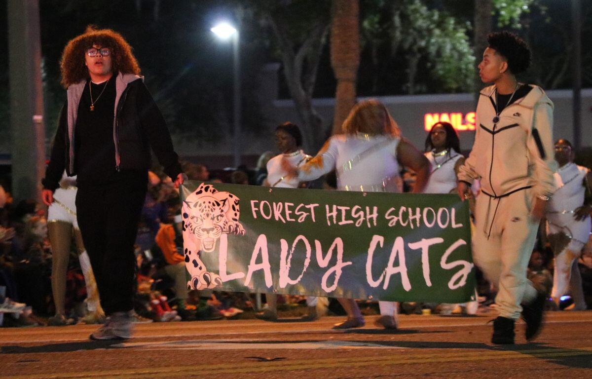 Two students hold a green sign with white text reading, "Forest High School Lady Cats." Girls in white uniforms dance in the background.