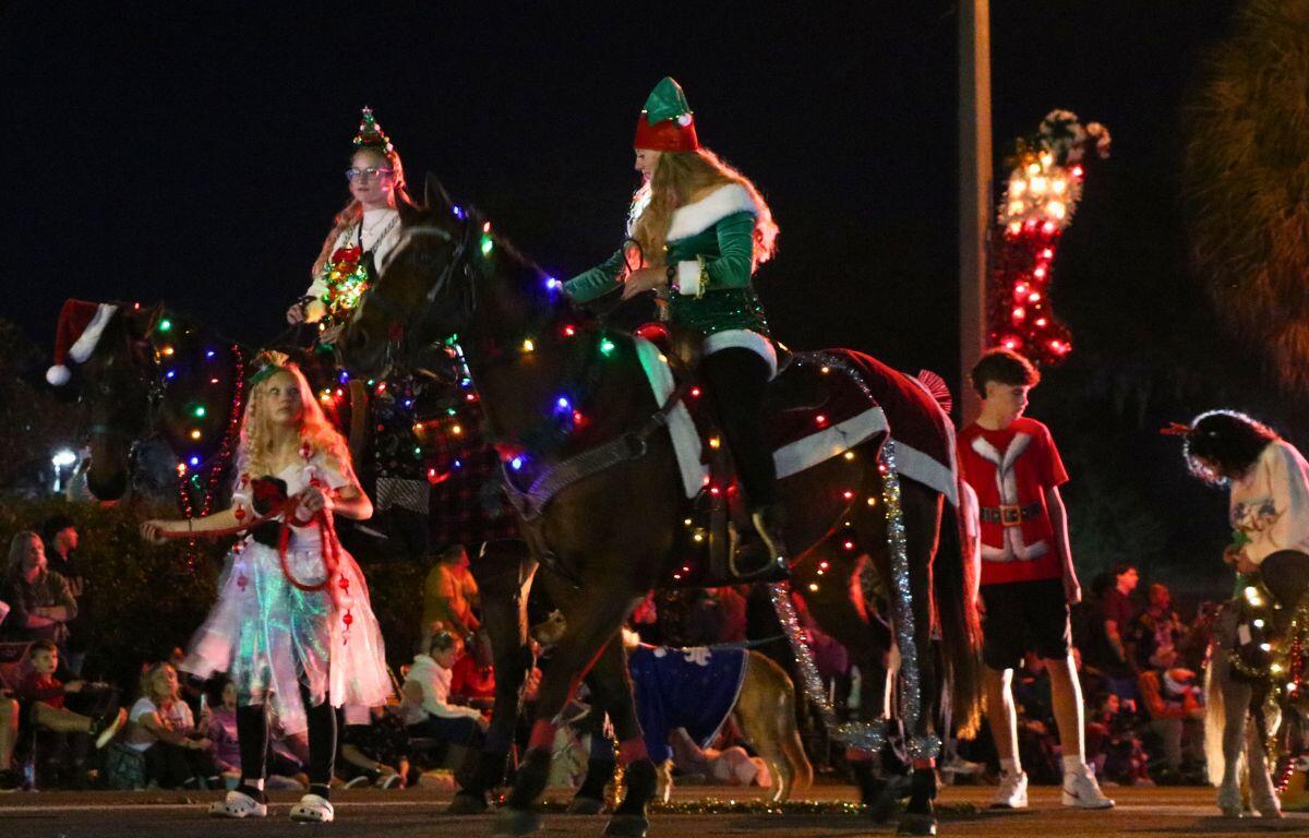 Two women sit on a horses with lights and decorations strewn on them. Kids surround them with a pony and wearing Christmas themed outfits.