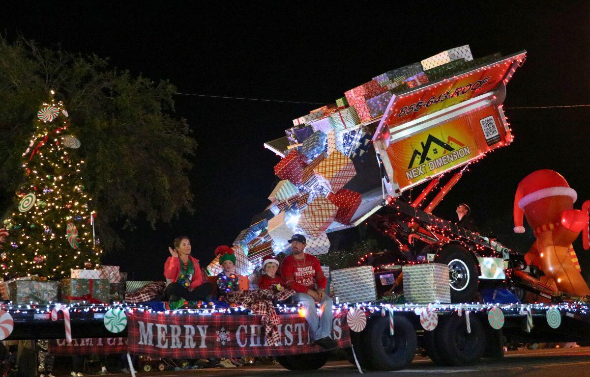 A large parade float features a decorative container unloading Christmas presents into the bed of a truck with inflatables and lights surrounding it.