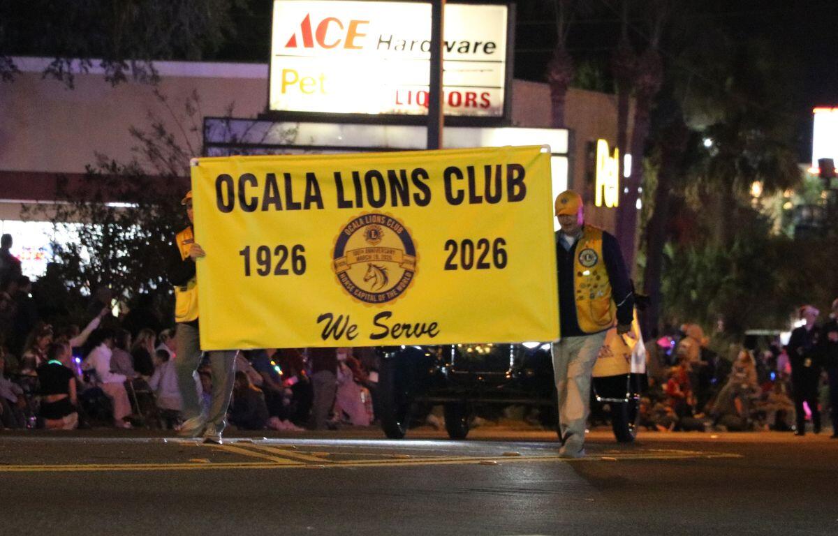 Two men in yellow vests hold a yellow sign reading, "Ocala Lions Club," as they walk down a street.