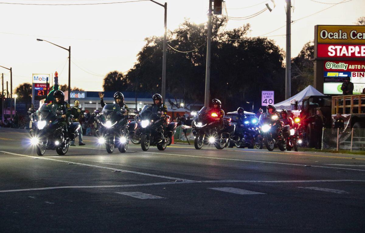 Cops on motorcycles drive down a road while waving to a crowd of people.