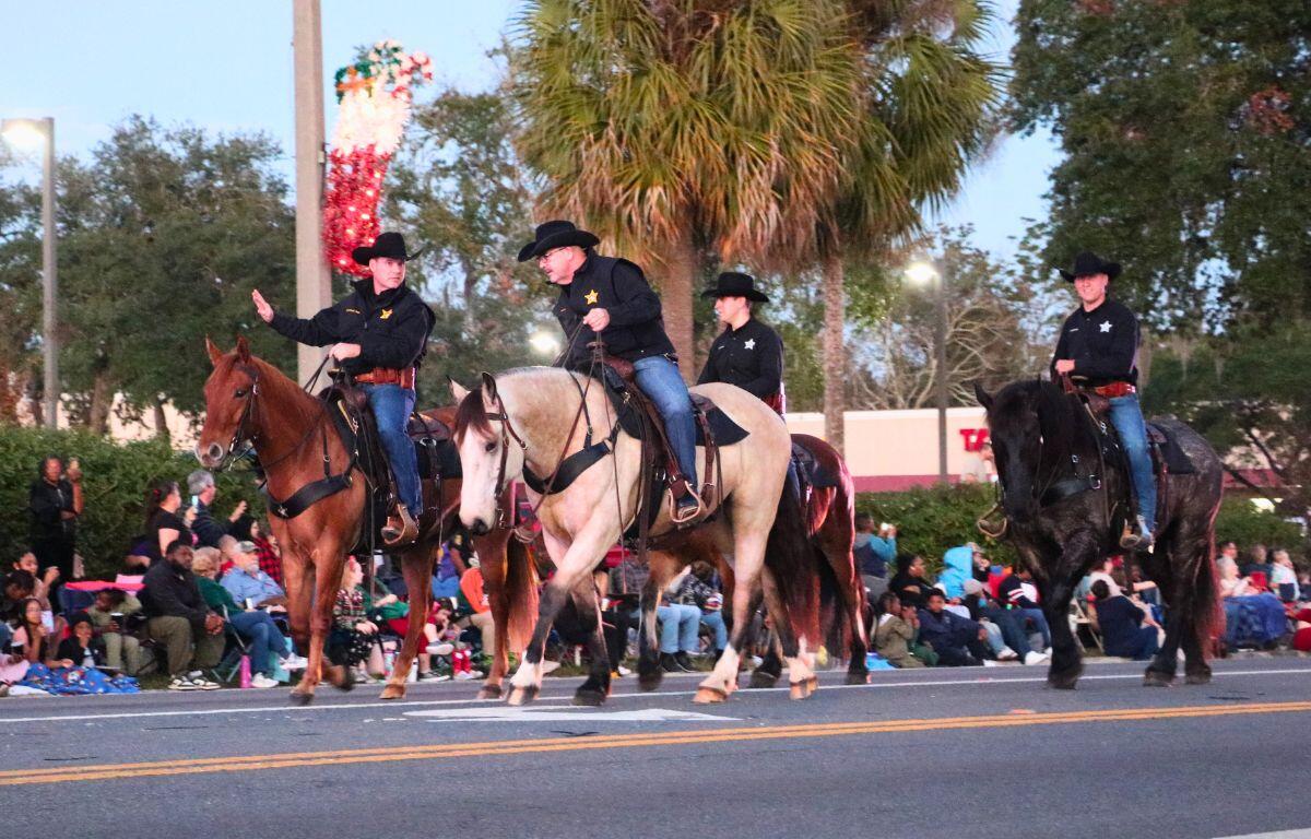 Police on horses parade together down a street while talking to each other and waving at a crowd in the background.