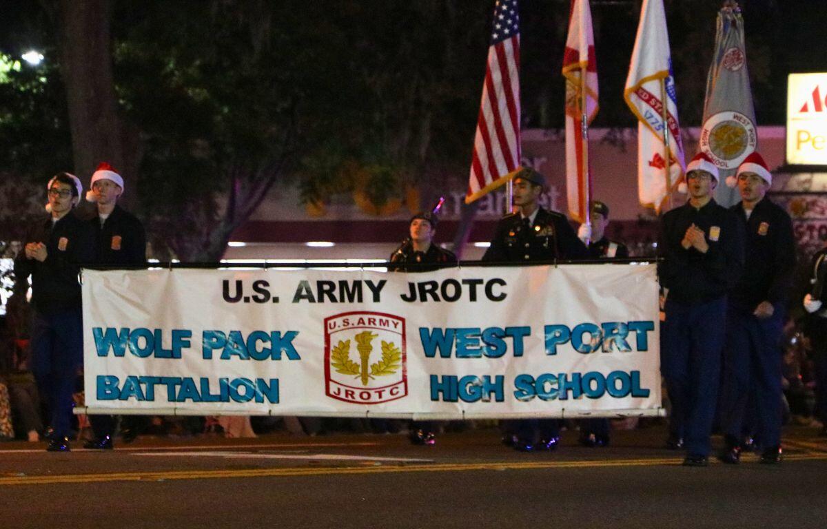 The West Port JROTC wear their uniforms with Christmas hats while holding banners and flags, walking down a dimly lit road.