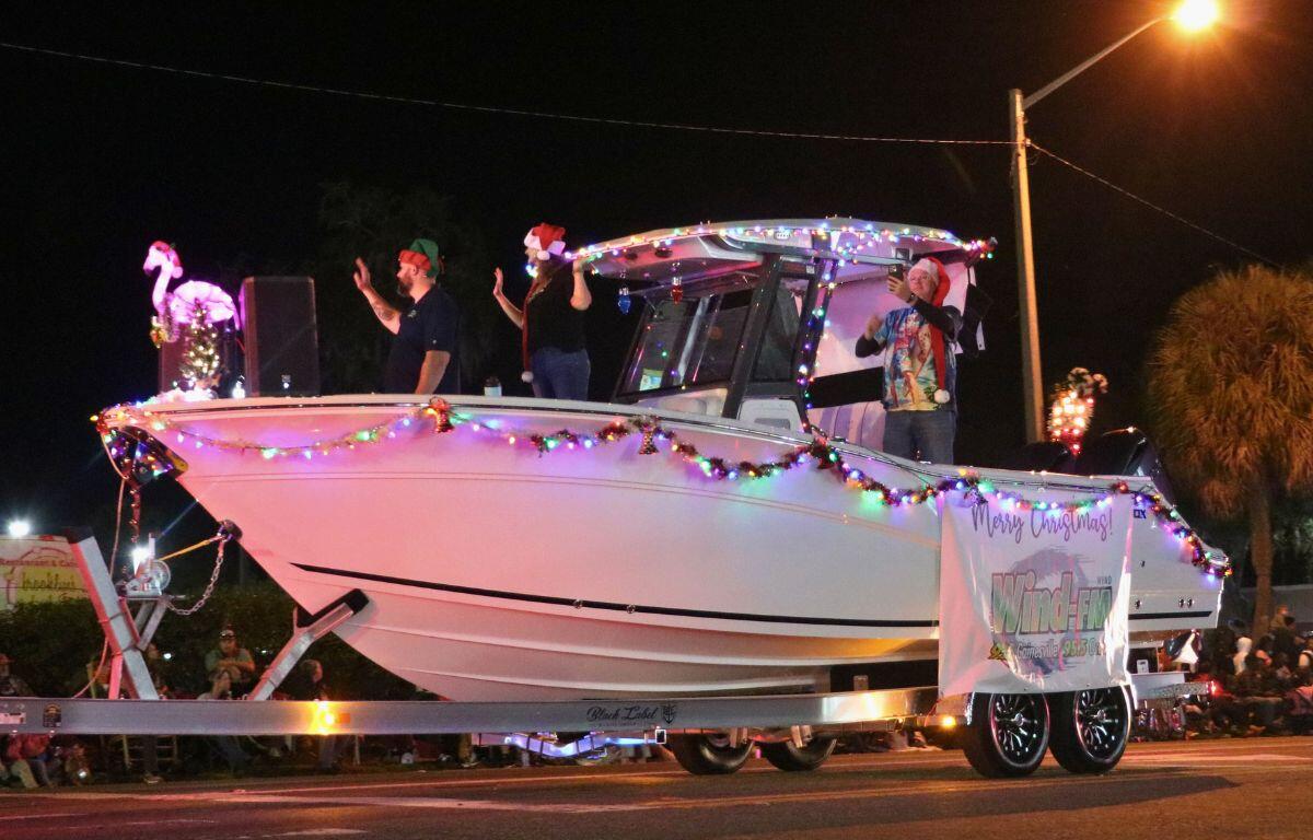 A group of people in festive Christmas hats stand together on a boat with Christmas lights hanging from it.