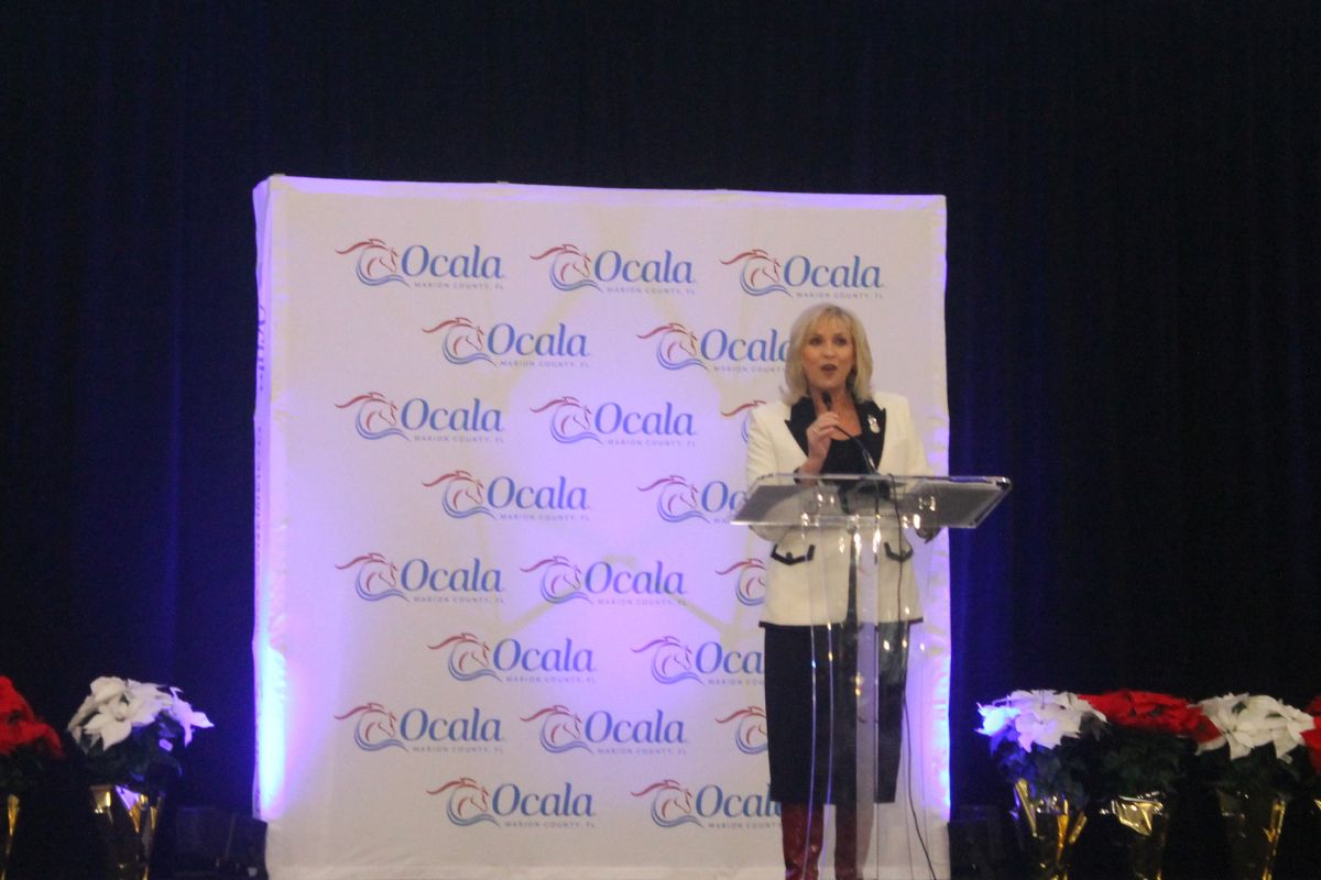 A well-dressed woman standing behind an acrylic microphone stand, holding a microphone in front of a huge white placard with writing, with a black background and potted plants to her left and right.