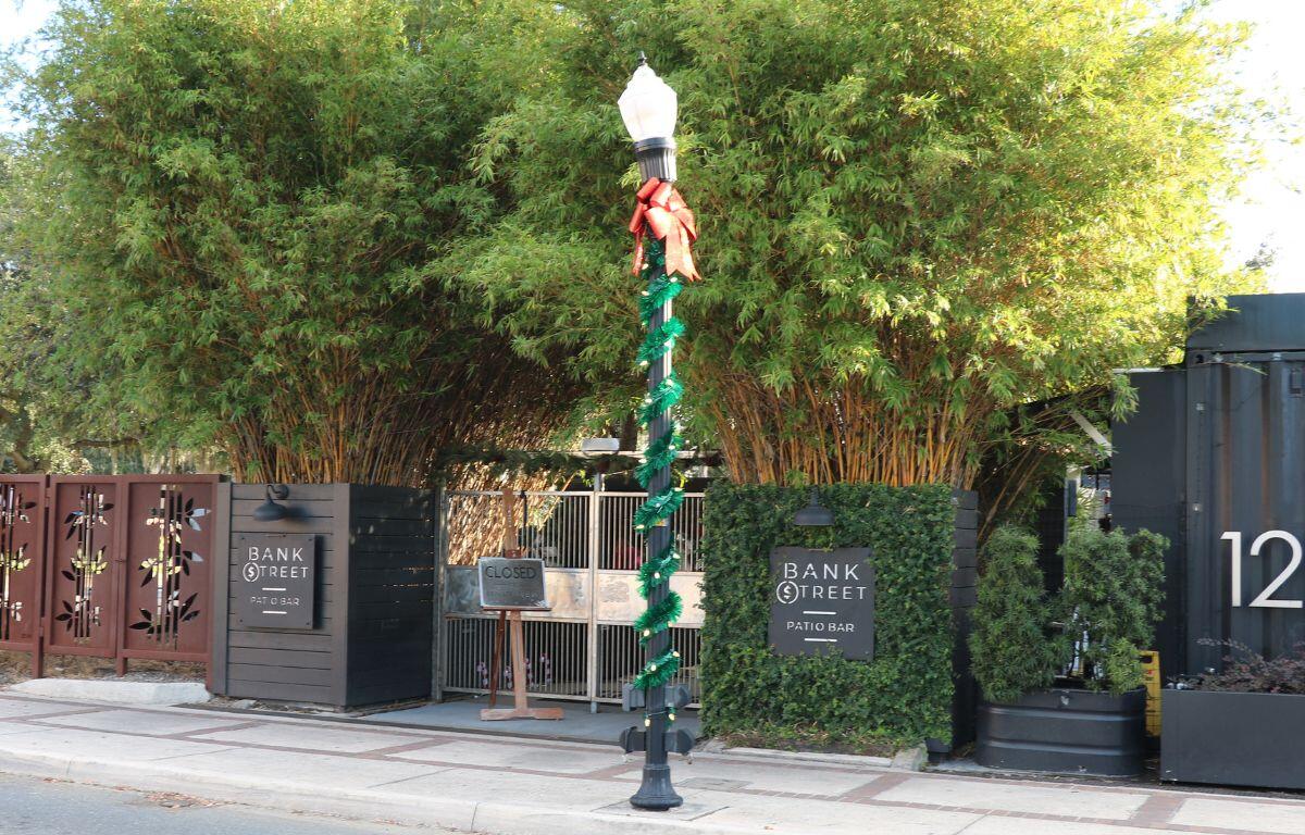 The exterior of a restaurant is fenced off with bushes surrounding it on the side of a road. Multiple signs read, "Bank Street Patio Bar" and "Closed."