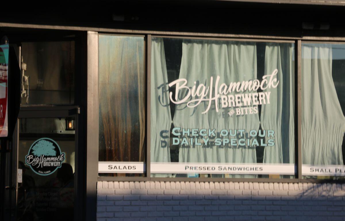 A restaurant front has white bricks below multiple large windows. A window and the door have signs reading, "Big Hammock Brewery."