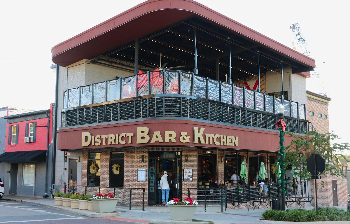 A large restaurant front has two stories with the top story being an open-air patio. The first floor has a brick exterior with a large, red strip separating the floors. The red strip has a sign reading, "District Bar & Kitchen."