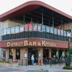 A large restaurant front has two stories with the top story being an open-air patio. The first floor has a brick exterior with a large, red strip separating the floors. The red strip has a sign reading, "District Bar & Kitchen."
