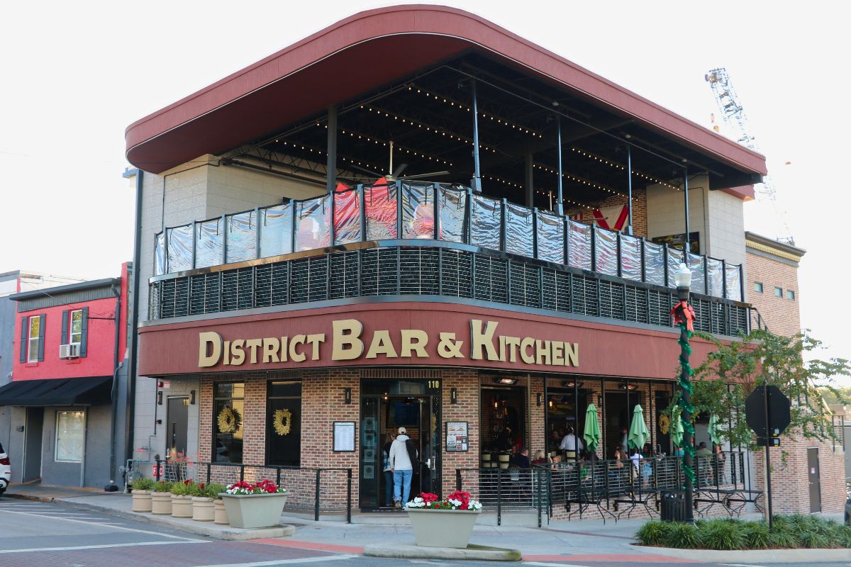A large restaurant front has two stories with the top story being an open-air patio. The first floor has a brick exterior with a large, red strip separating the floors. The red strip has a sign reading, "District Bar & Kitchen."