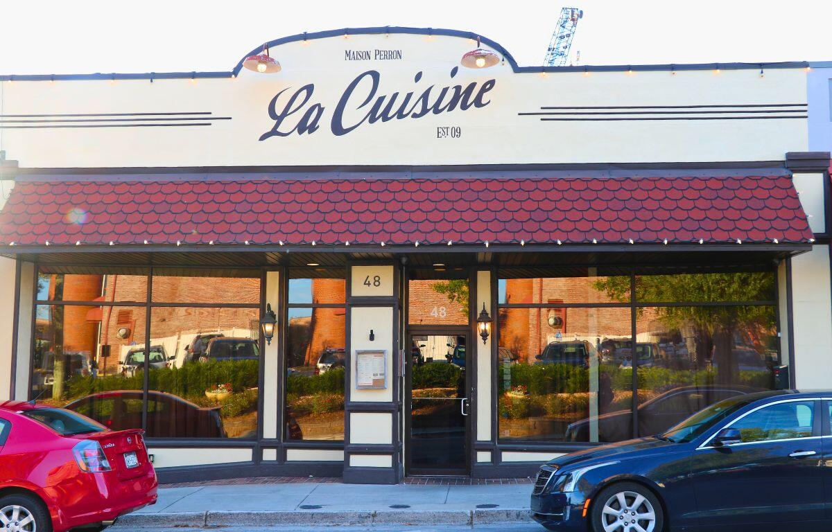 A restaurant front has a beige exterior with red panels above the entrance. Two cars are parallel parked in front of the restaurant. "La Cuisine" is written above.