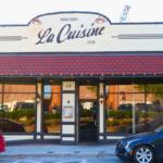A restaurant front has a beige exterior with red panels above the entrance. Two cars are parallel parked in front of the restaurant. "La Cuisine" is written above.