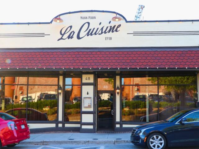 A restaurant front has a beige exterior with red panels above the entrance. Two cars are parallel parked in front of the restaurant. "La Cuisine" is written above.