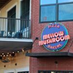 A brick front restaurant with a balcony has a large blue and red sign reading, " Pizza. Mellow Mushroom. Bakers."