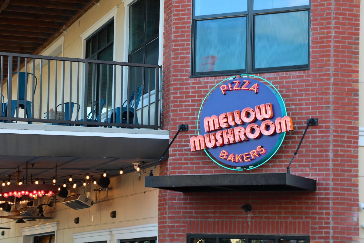 A brick front restaurant with a balcony has a large blue and red sign reading, " Pizza. Mellow Mushroom. Bakers."
