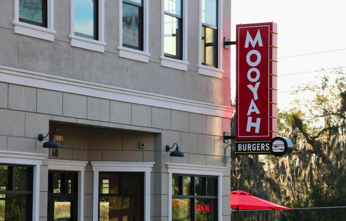 A restaurant front is made from gray bricks with lights and a camera above the entrance. Hanging from the building is a red. white and black sign reading, "Mooyah Burgers."
