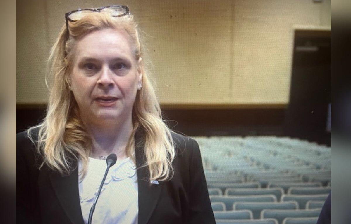 A well-dressed woman standing in front of a microphone, with sears to her right and in the background. In the far background, gray seats, brown baseboards and off-beige walls.
