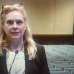 A well-dressed woman standing in front of a microphone, with sears to her right and in the background. In the far background, gray seats, brown baseboards and off-beige walls.