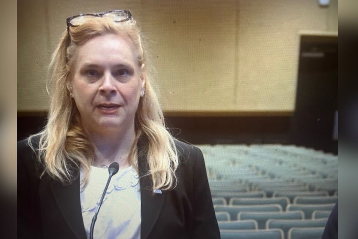 A well-dressed woman standing in front of a microphone, with sears to her right and in the background. In the far background, gray seats, brown baseboards and off-beige walls.