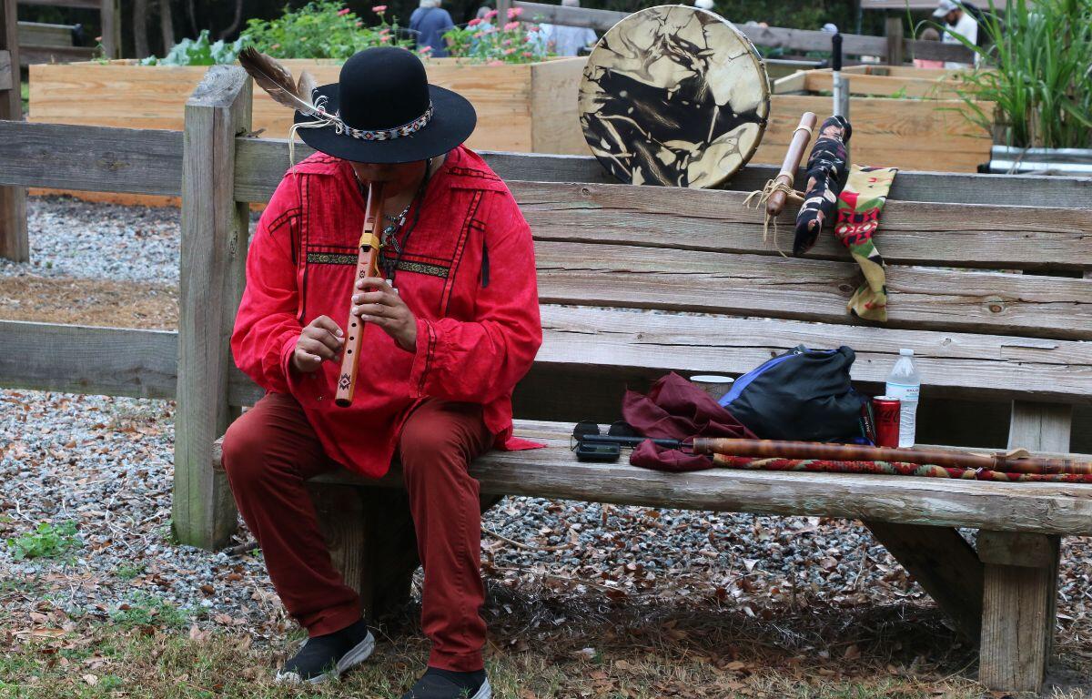 A man dressed as a Seminole plays flute on a bench in front of a garden.