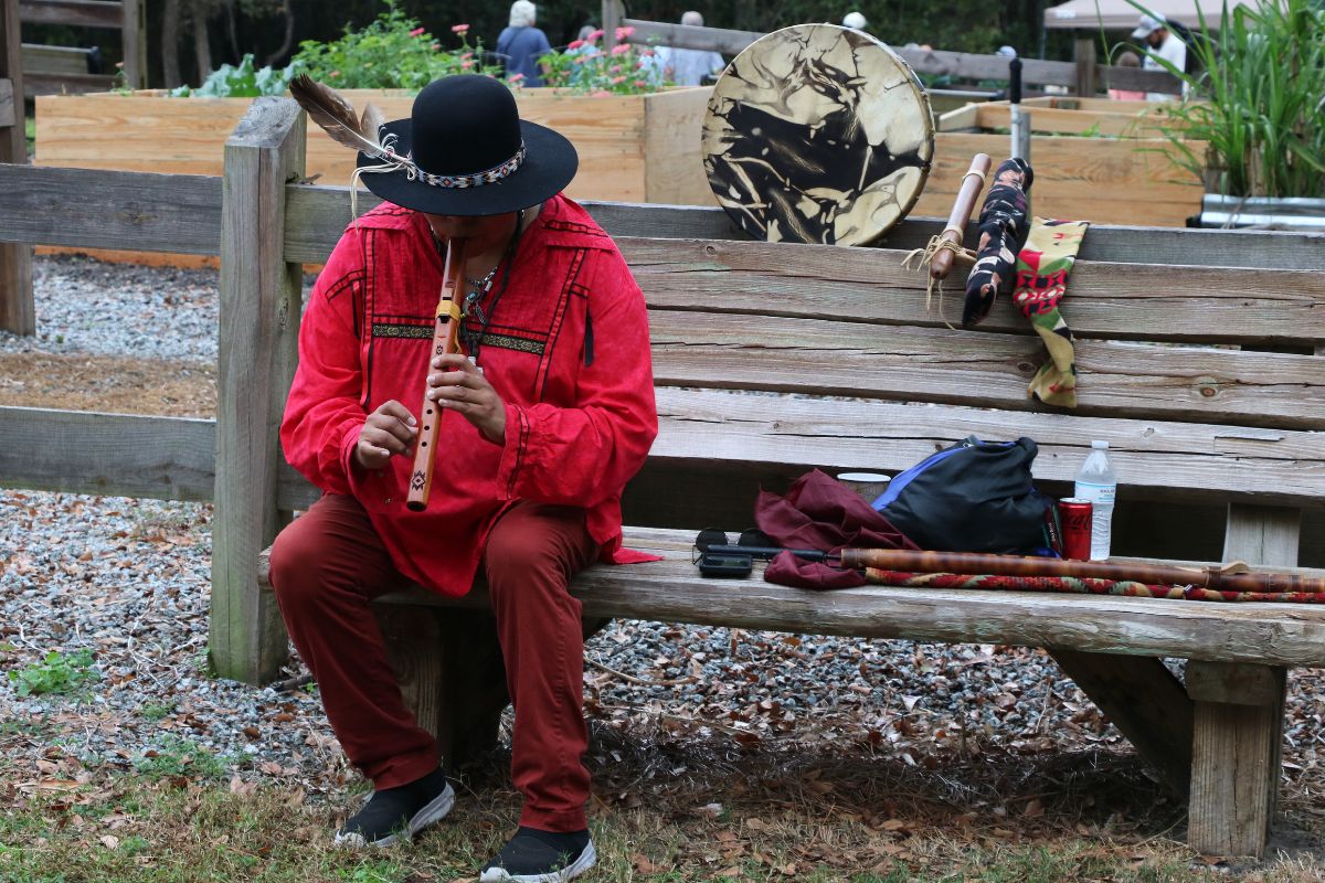 A man dressed as a Seminole plays flute on a bench in front of a garden.