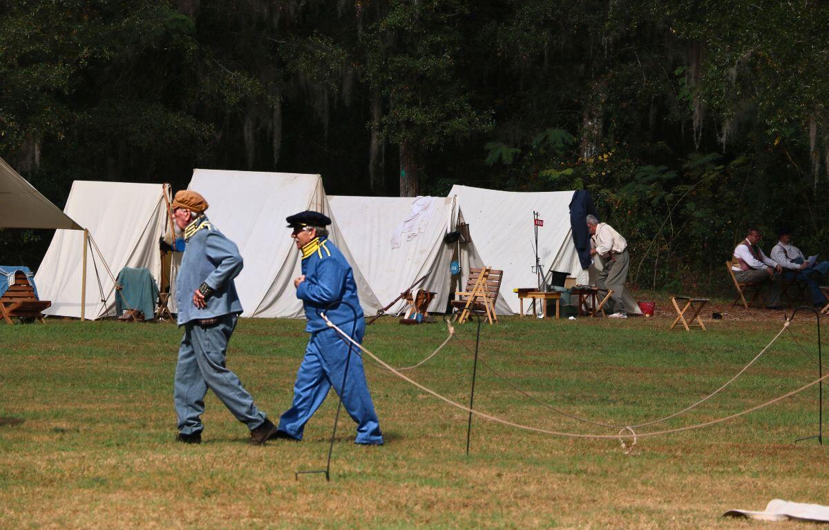 Two men in historical soldier uniforms walk through a field surrounded by a dense forest with white tents pitched around the area.