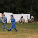 Two men in historical soldier uniforms walk through a field surrounded by a dense forest with white tents pitched around the area.
