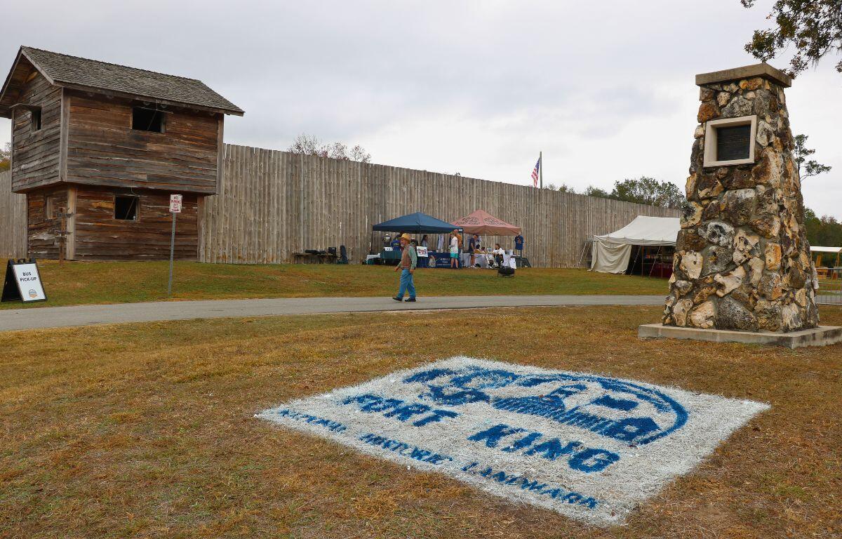 A large fort stands in the background with an American flag stretching over its wall and two vendor booths lined in front of it. On the ground across a field from the fort is a painted white square with a blue drawing of the fort and words reading, "Fort King National Historic Landmark." A tall pillar stands next to it as people walk around the area.