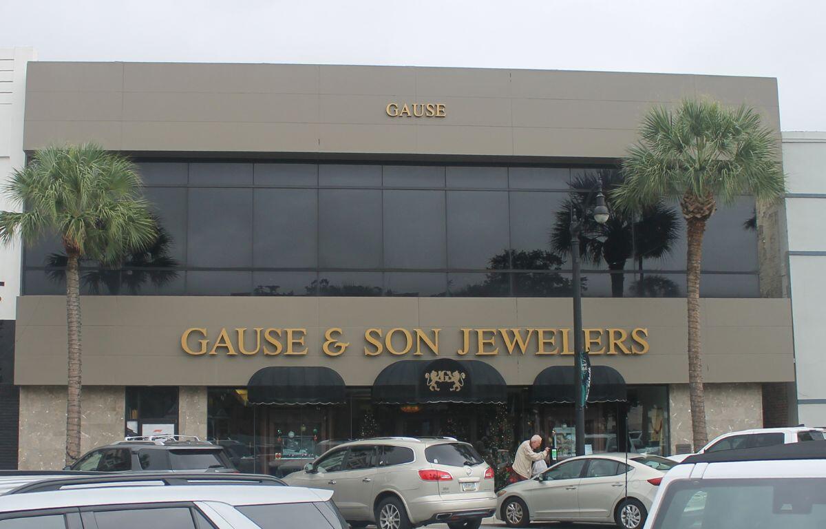 Background gray and blue sky, palm to the left and right of building, building with gray face and many windows on second floor and lettering on front of building. Canopy over front door. Cars, human and parking meters in the foreground.