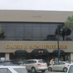 Background gray and blue sky, palm to the left and right of building, building with gray face and many windows on second floor and lettering on front of building. Canopy over front door. Cars, human and parking meters in the foreground.