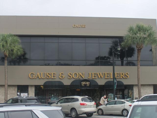 Background gray and blue sky, palm to the left and right of building, building with gray face and many windows on second floor and lettering on front of building. Canopy over front door. Cars, human and parking meters in the foreground.