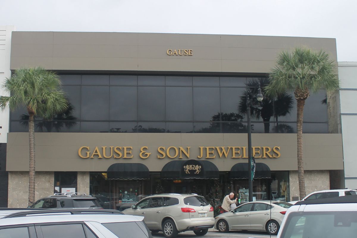 Background gray and blue sky, palm to the left and right of building, building with gray face and many windows on second floor and lettering on front of building. Canopy over front door. Cars, human and parking meters in the foreground.