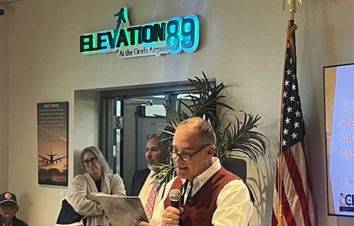 in the background on the left a man wearing a ball cap, a woman and man standing against the wall, with a neon sign over their head, and a plastic plant and an American flag to their right. With a man reading from a document holding a microphone in the foreground.