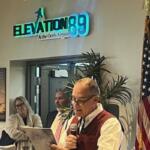 in the background on the left a man wearing a ball cap, a woman and man standing against the wall, with a neon sign over their head, and a plastic plant and an American flag to their right. With a man reading from a document holding a microphone in the foreground.