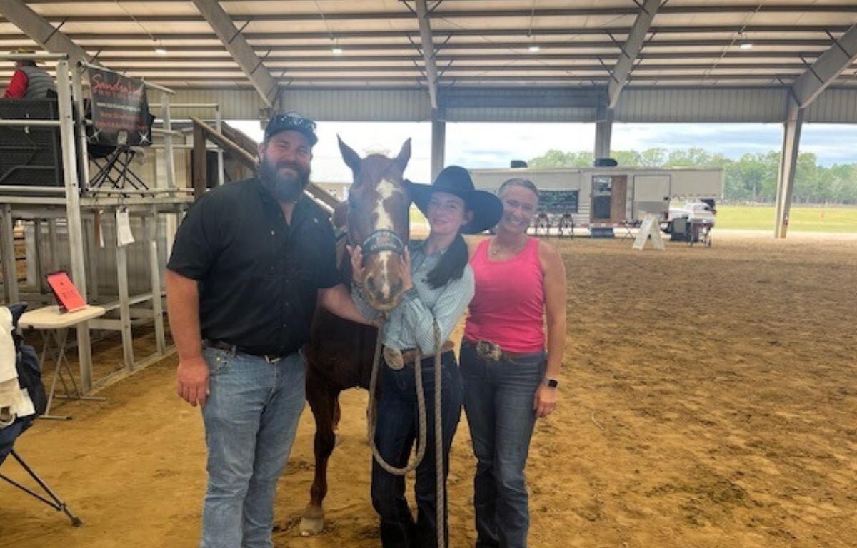 Chairs and metal riser stands with banner on the left, in the background, a roof, RV on a clay surface, with a man in a baseball hat, a horse, young woman wearing a cowboy hat, and woman wearing a pink top on the right.