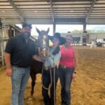 Chairs and metal riser stands with banner on the left, in the background, a roof, RV on a clay surface, with a man in a baseball hat, a horse, young woman wearing a cowboy hat, and woman wearing a pink top on the right.