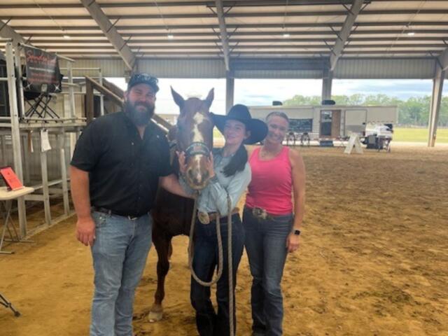 Chairs and metal riser stands with banner on the left, in the background, a roof, RV on a clay surface, with a man in a baseball hat, a horse, young woman wearing a cowboy hat, and woman wearing a pink top on the right.