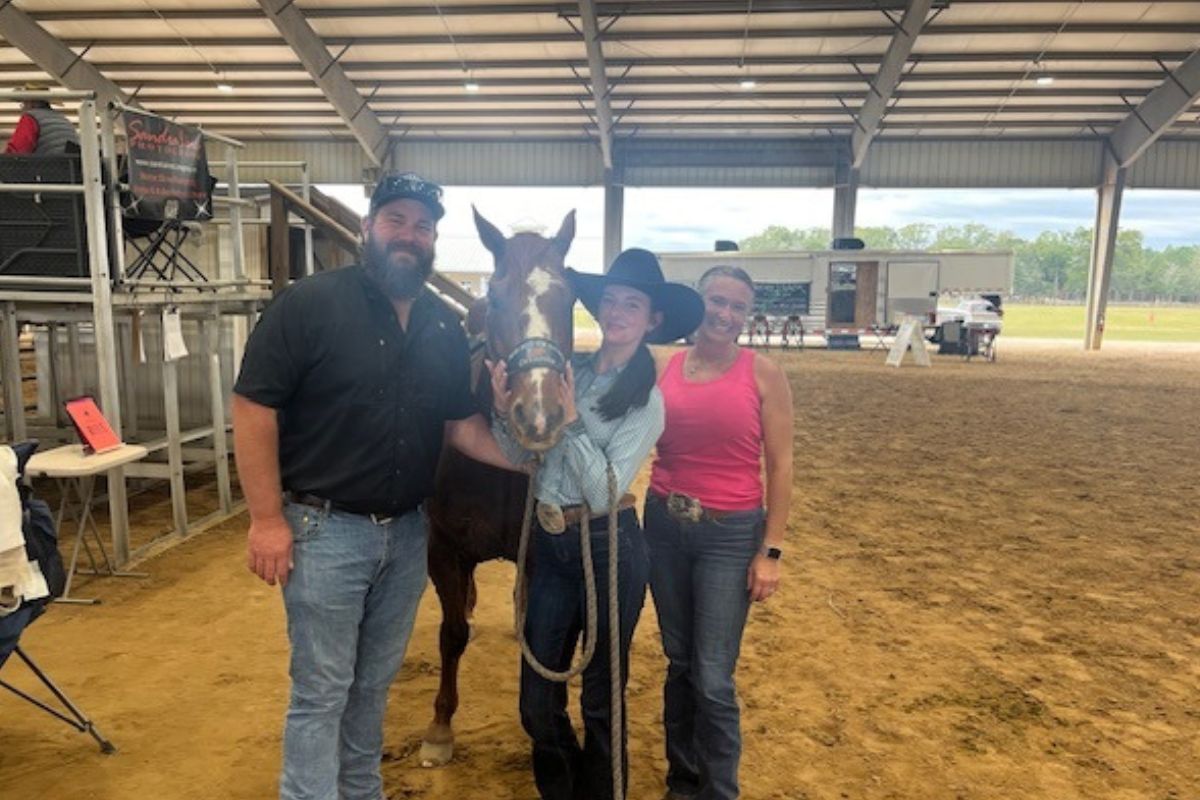 Chairs and metal riser stands with banner on the left, in the background, a roof, RV on a clay surface, with a man in a baseball hat, a horse, young woman wearing a cowboy hat, and woman wearing a pink top on the right.