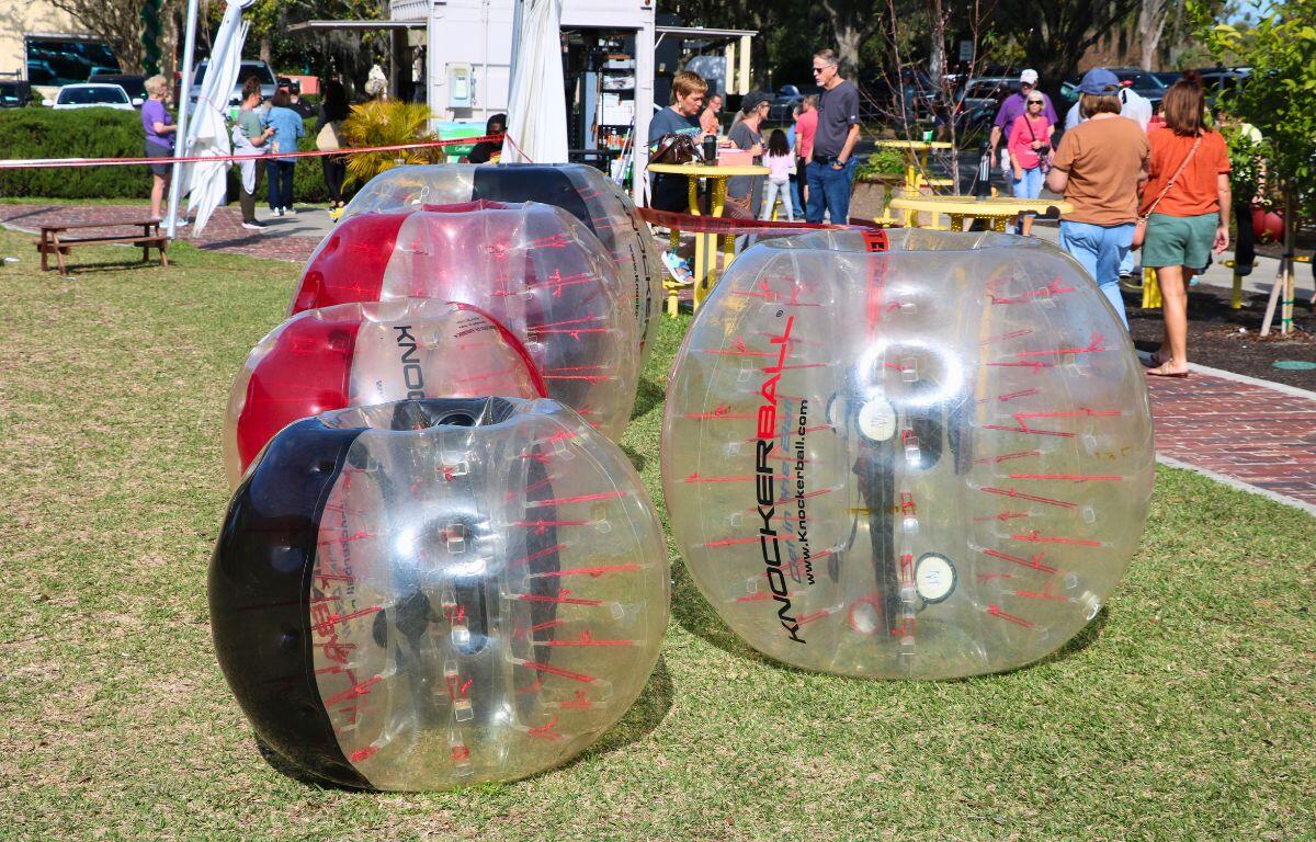 Human hamster balls sit in a patch of grass next to a sidewalk with yellow tables on it.