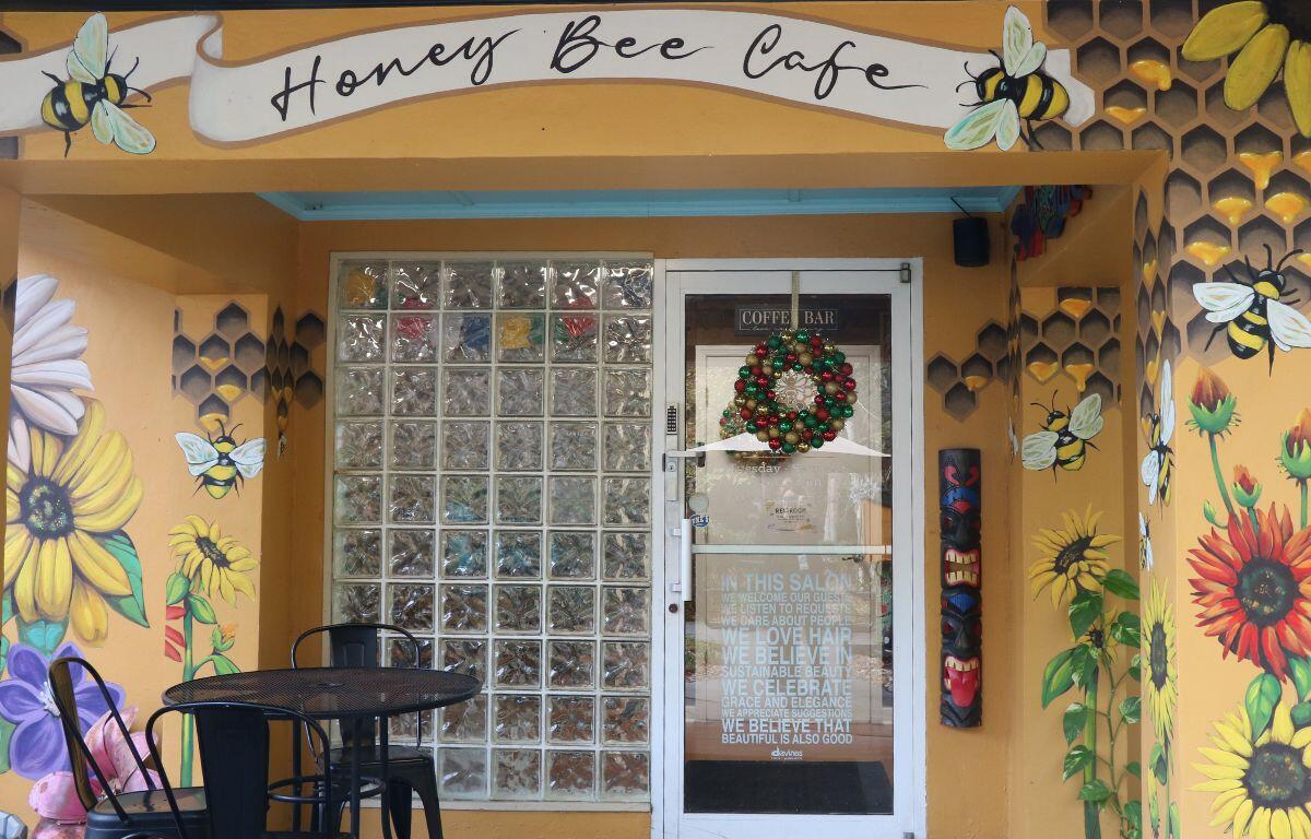 A café with a yellow exterior has flowers and bees painted over the entrance with black paint reading, "Honey Bee Cafe." The outer entrance features a glass window and door and a black table with chairs.