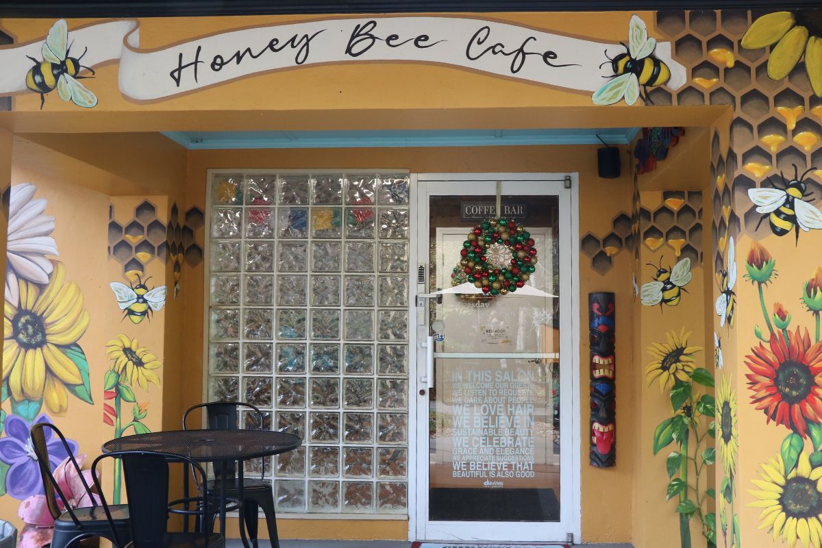 A café with a yellow exterior has flowers and bees painted over the entrance with black paint reading, "Honey Bee Cafe." The outer entrance features a glass window and door and a black table with chairs.
