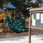 A board with flyers in it stands in playground with and open field, paved pathway and trees around it.