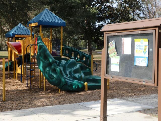 A board with flyers in it stands in playground with and open field, paved pathway and trees around it.