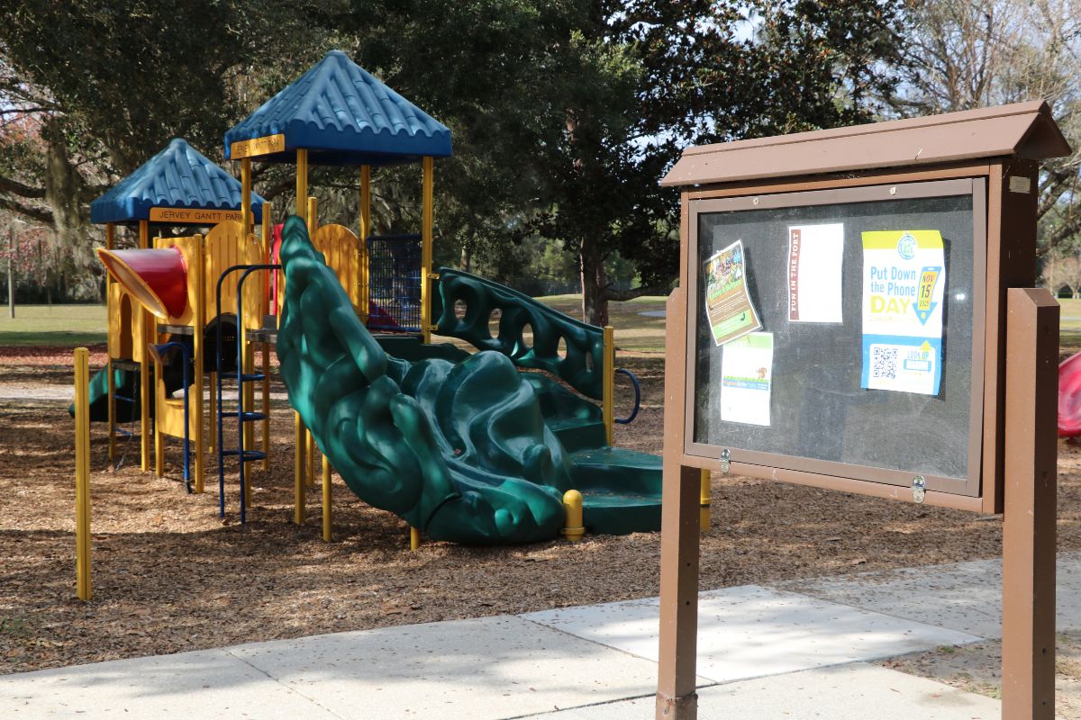 A board with flyers in it stands in playground with and open field, paved pathway and trees around it.